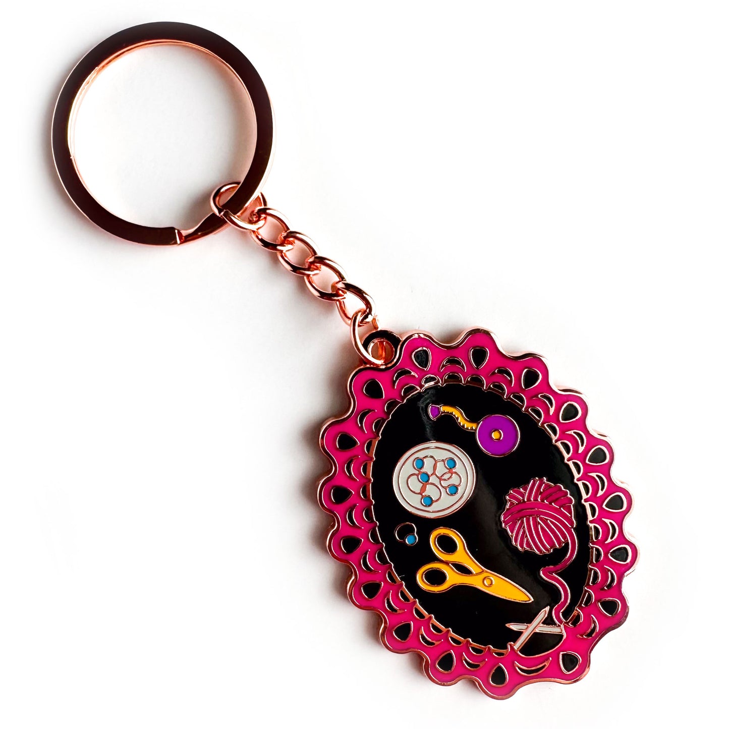 A copper metal color enamel keychain, the charm of the keychain is shaped like a lace knitting project still on the needles with a pink yarn ball, scissors, stitch markers, and a tape measure. 