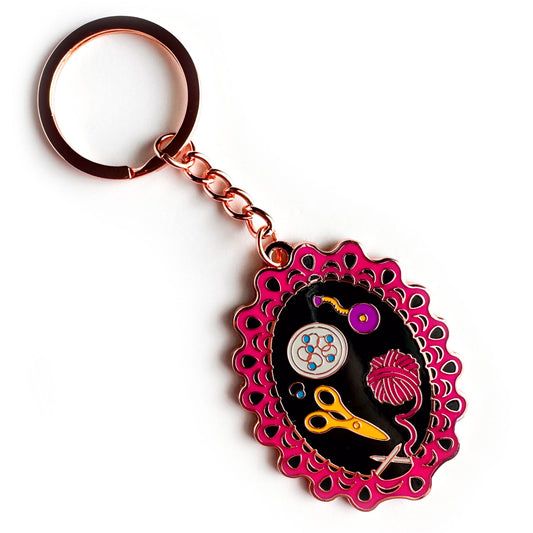 A copper metal color enamel keychain, the charm of the keychain is shaped like a lace knitting project still on the needles with a pink yarn ball, scissors, stitch markers, and a tape measure.