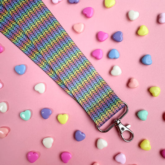 A woven lanyard with a pastel rainbow stockinette stitch graphic printed on it with a silver metal lobster claw clasp at the bottom. The lanyard is on a pastel pink background with pastel colored heart beads scattered around it.