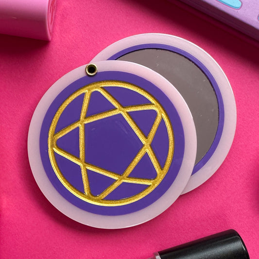 A circular hanged compact mirror that is a purple circle with a pastel pink border with a gold shiny pentacle embossed on the cover.  The mirror is on a hot pink background with some makeup around it. 