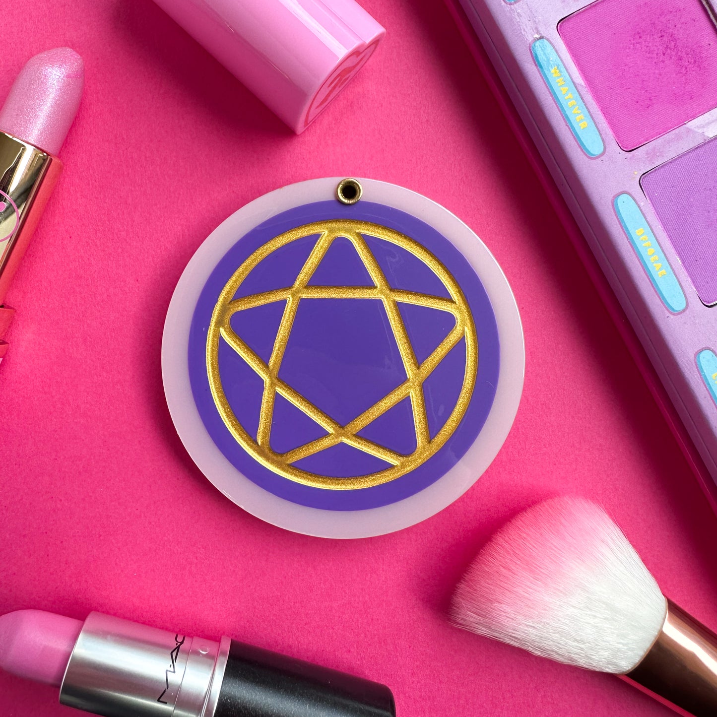 An acetate circle that is purple with a pastel pink border with an embossed gold pentacle on it. This item is on a hot pink background surrounded by some makeup