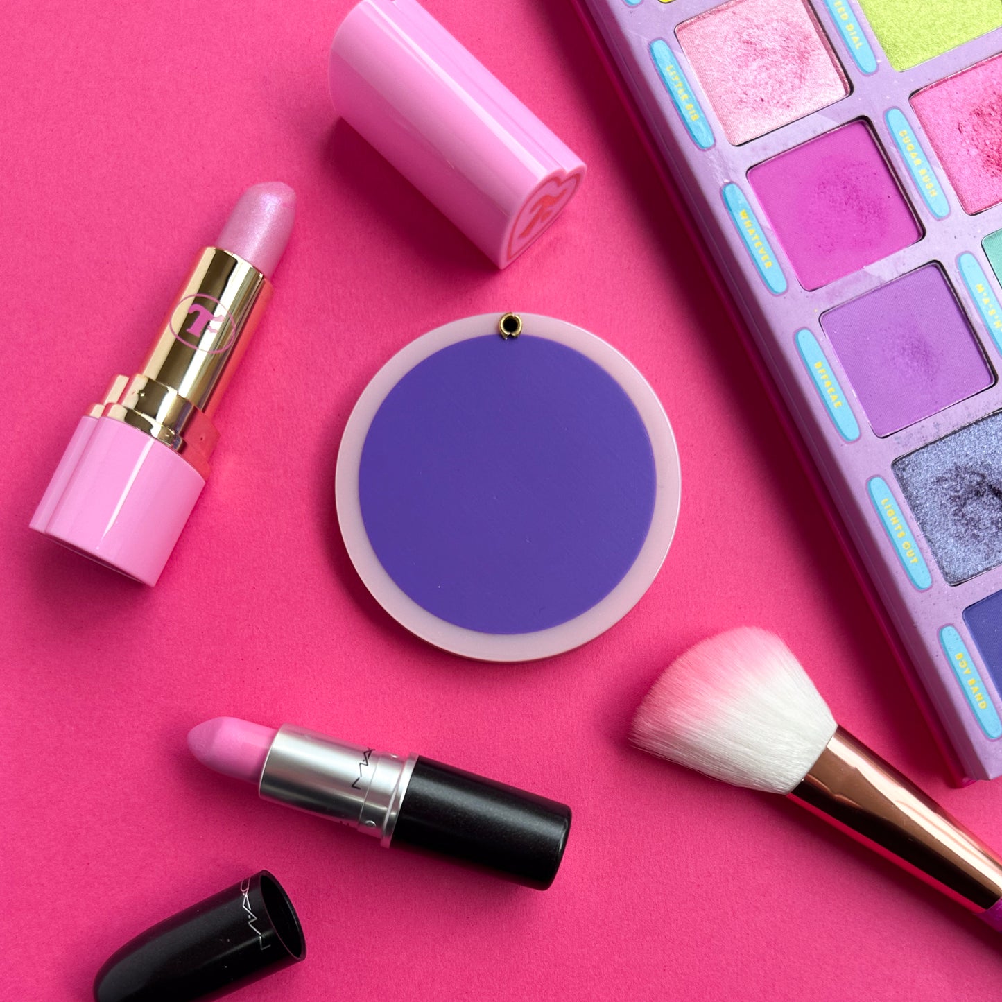 The back of the compact mirror that is a purple circle with a pastel pink border. The item is on a hot pink background and is surrounded by two open lip sticks, a pink makeup brush and a pink and purple eyeshadow palette. 