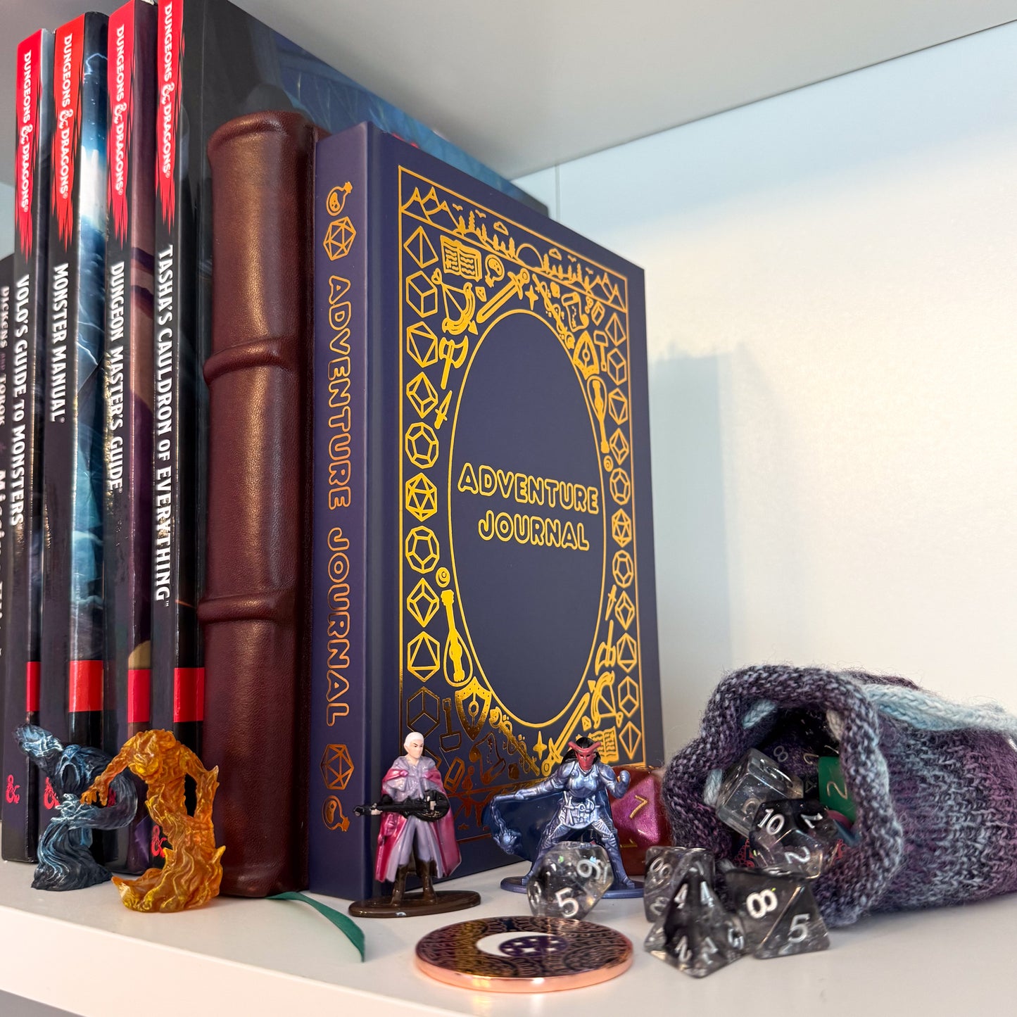 The Adventure Journal displayed on a bookshelf with Dungeons and Dragons guides, figurines, and an overflowing dice bag.