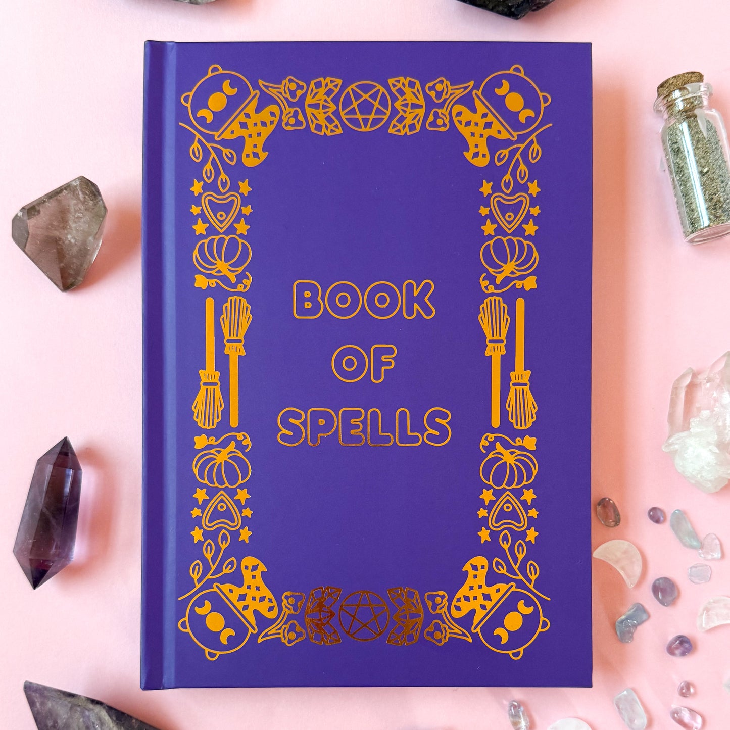 The Bool of Spells on a light pink background surrounded by crystals and spell jars.