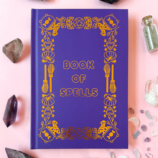 The Bool of Spells on a light pink background surrounded by crystals and spell jars.