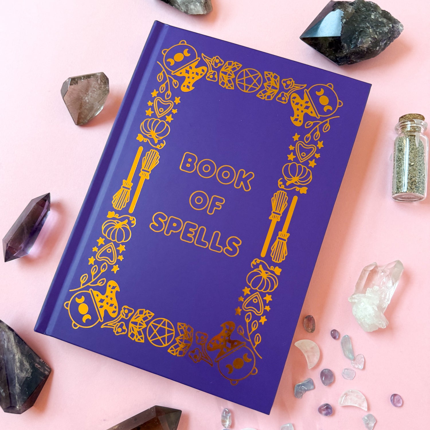 The Bool of Spells on a light pink background surrounded by crystals and spell jars.