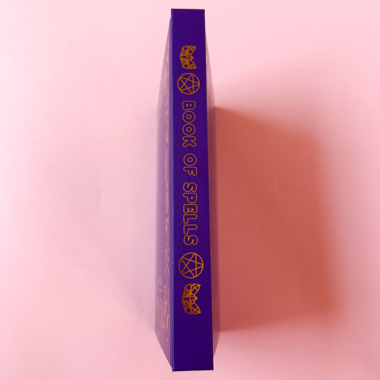 The spine of the Book of Spells with the title in bubble letters surrounded by illustrations of crystals and pentacles. All details are in gold foil.