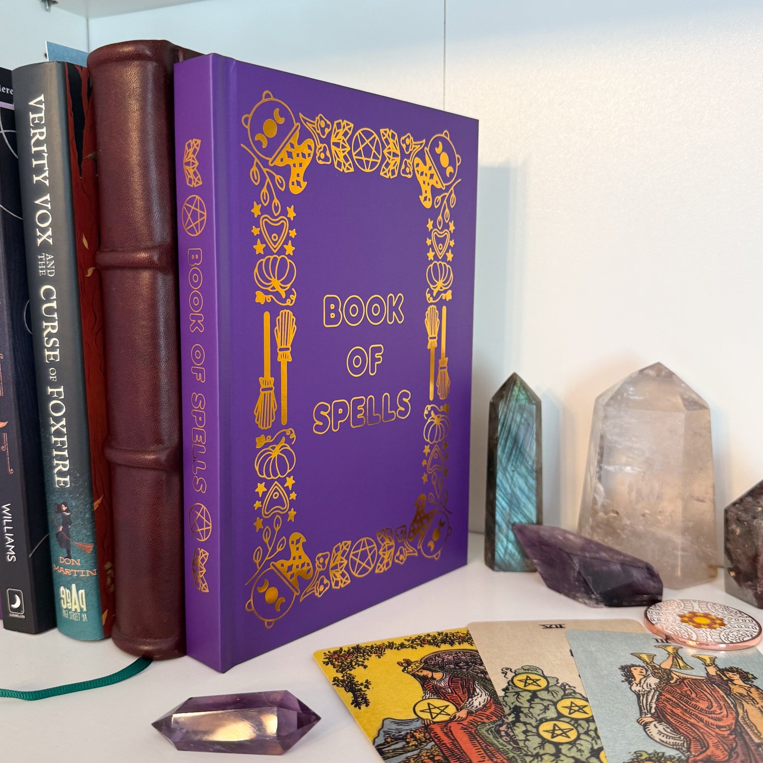 The Book of Spells displayed on a shelf with other books, crystals, tarot cards, and a divination coin. 