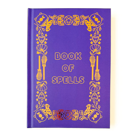 A purple hard covered notebook with gold foil detailing. The title Book of Spells is in the center, and the edges are decorated in witchy illustrations. A few examples of these illustrations are cauldrons, crystals, brooms, and pentacles.   