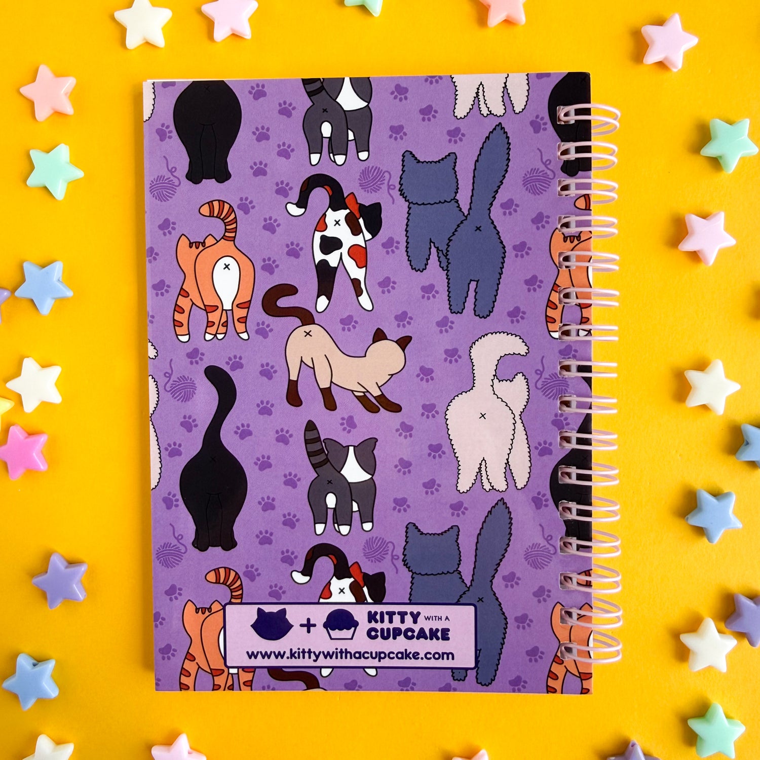 A spiral bound notebook with a purple cover and pattern of different cat butts on it. There is a the Kitty With A Cupcake logo on the bottom. The notebook is on a yellow background with plastic star beads around it. 
