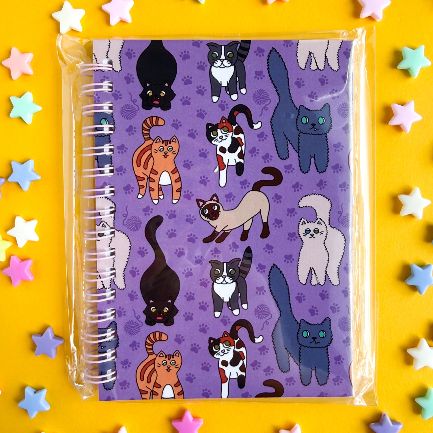 A purple spiral notebook with cute illustrations of cats on it wrapped in a clear plastic bag. The notebook is on a yellow background with plastic star beads around it. 