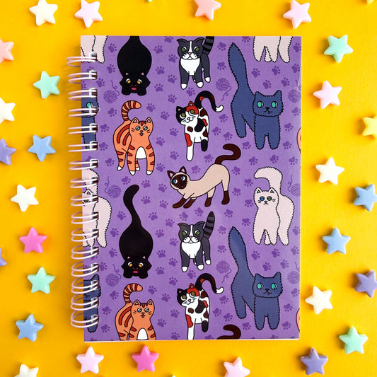 A spiral bound notebook with a purple cover and pattern of different cartoon cat illustrations on it, there are orange cats, calicos, siamese, a white fluffy cat, a Russian blue, a grey tabby, and a black cat. The notebook is on a yellow background with purple plastic star beads around it.
