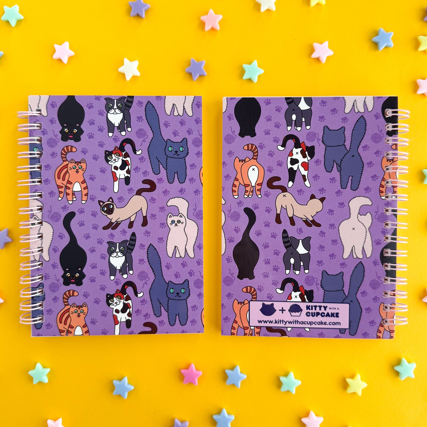 Two of the same notebook side by side showing the front and back cover of the notebook. The front cover has a purple background and features illustrations of different cats. The back cover has the same purple background but features illustrations of the behinds of the same cats. 