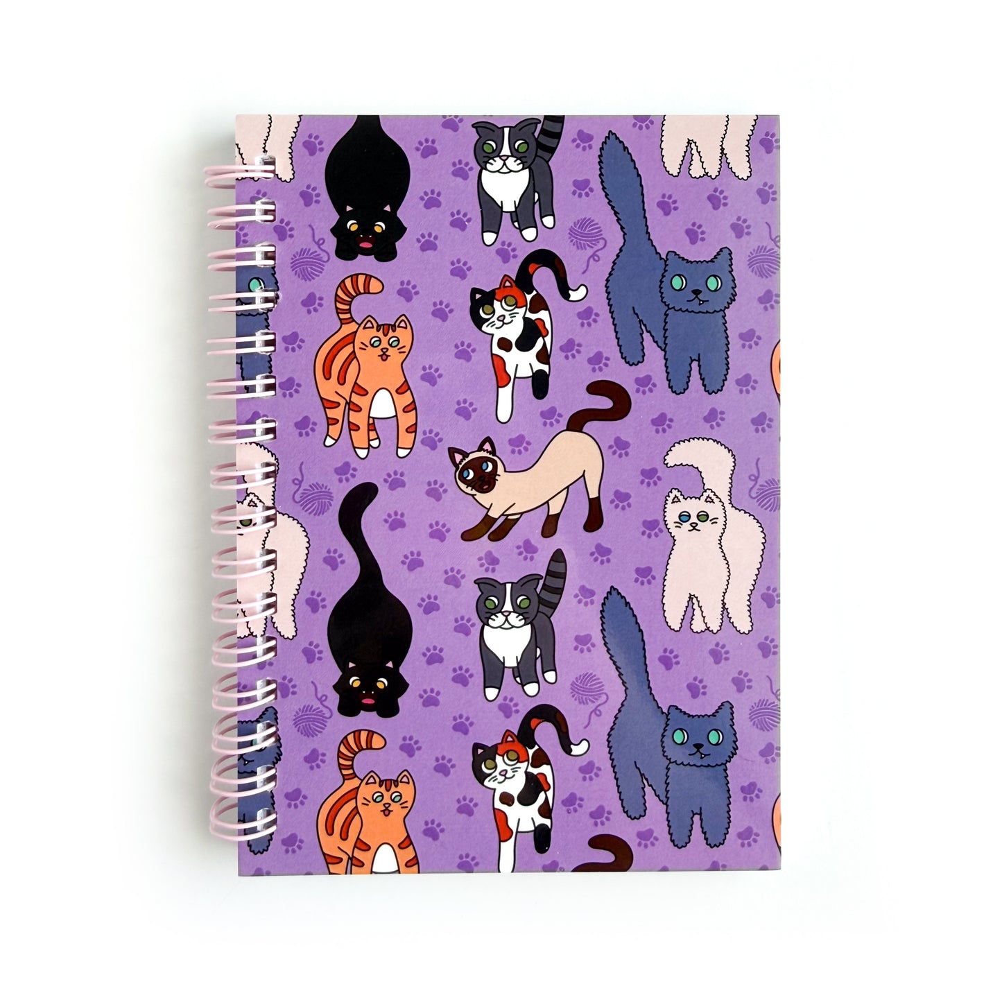 A spiral bound notebook with a purple cover and pattern of different cartoon cat illustrations on it. 