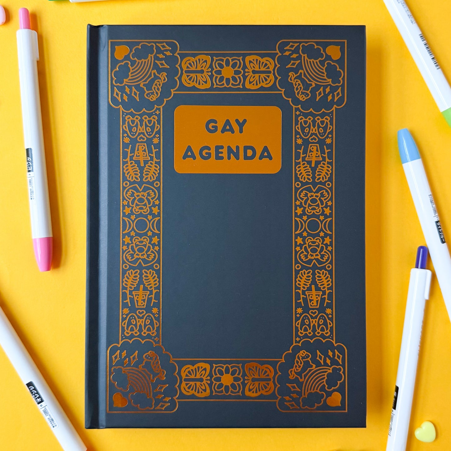 The Gay Agenda on a yellow background surrounded by colorful pens.