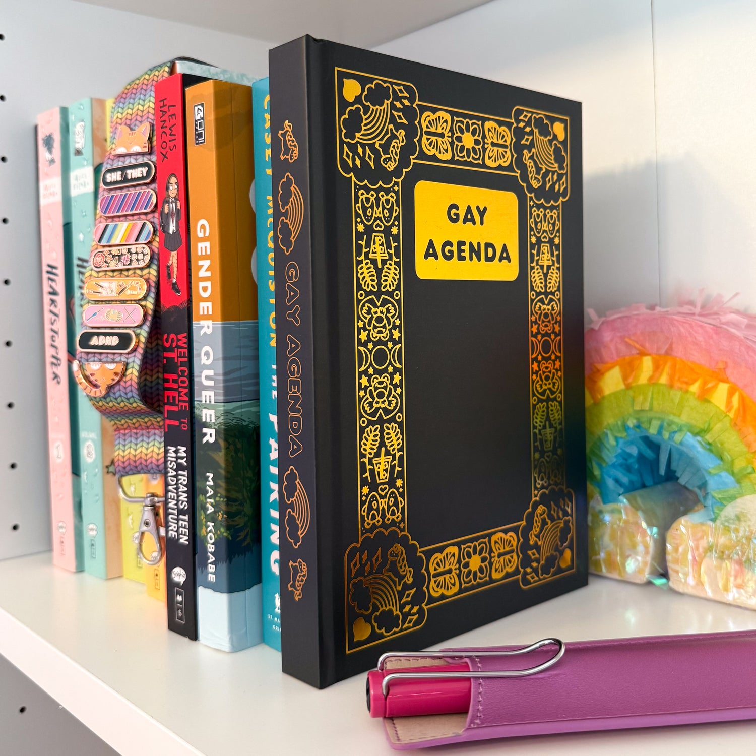 The Gay Agenda displayed on a bookshelf with other LGBTQ+ books, pride pals, a pen, and a pastel rainbow decoration.