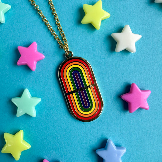 A gold necklace with a gold oval pendant with mirrored rainbows on it. The necklace is on a blue background with plastic star beads around it. 