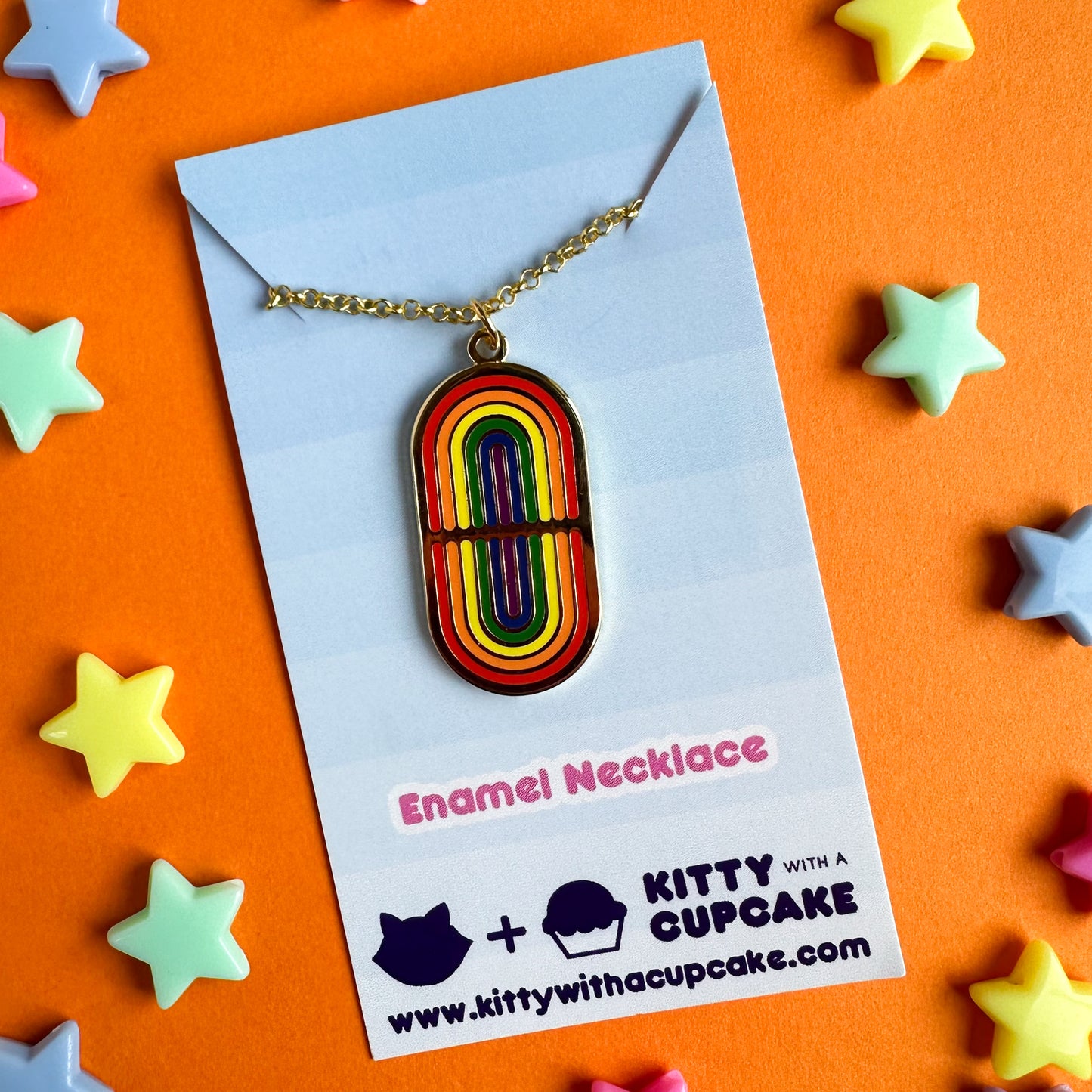 A gold pendant style necklace with an oval pendant with mirrored rainbows on it. The necklace is packaged on a blue card. The card is on an orange background with plastic stars around it. 