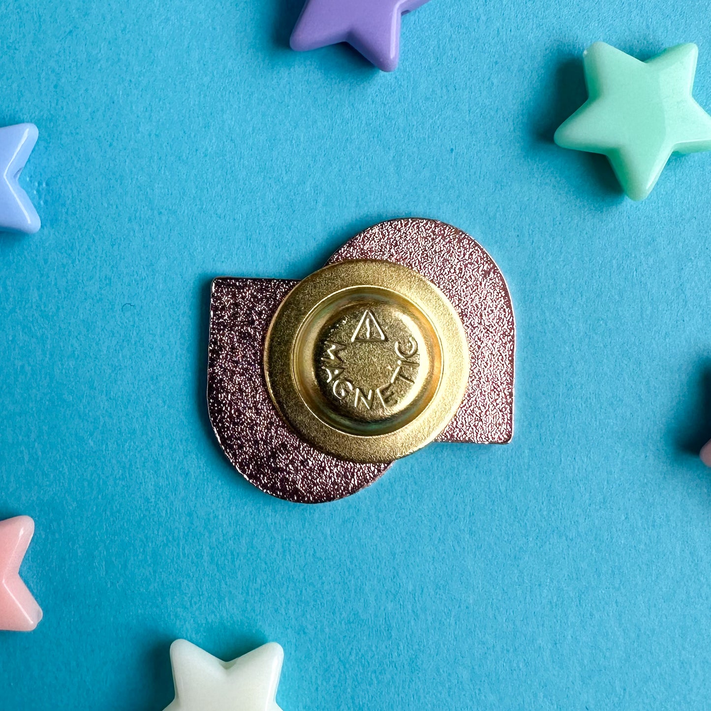 The underside of the rainbow squiggle pin with its magnetic back closure. The pin is on a blue paper background with plastic star beads around it. 