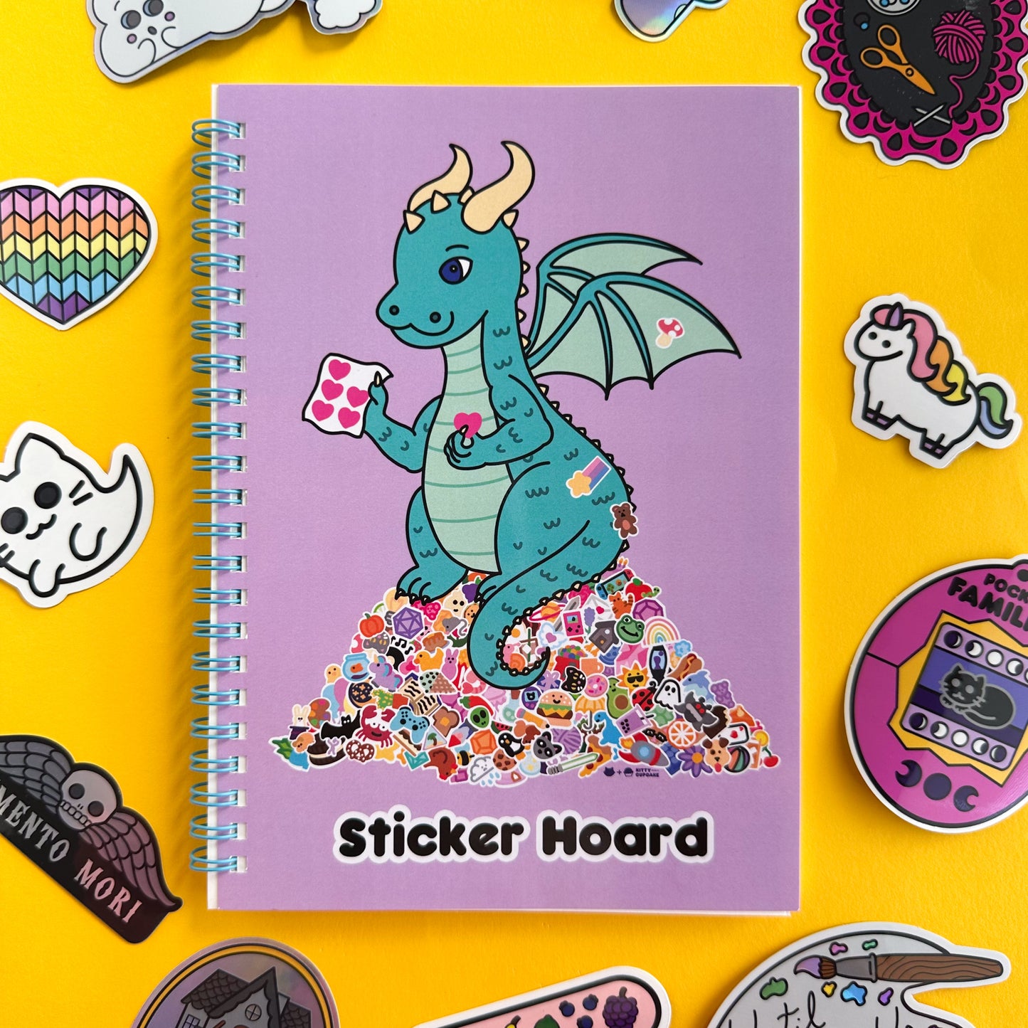 A spiral bound notebook with a lavender cover with an illustration of a teal dragon holding stickers and sitting on top of a giant pile of stickers. Underneath the dragon are the words "Sticker hoard" The notebook is sitting on a yellow piece of paper surrounded by cute vinyl stickers in the same style as the illustration of the dragon.