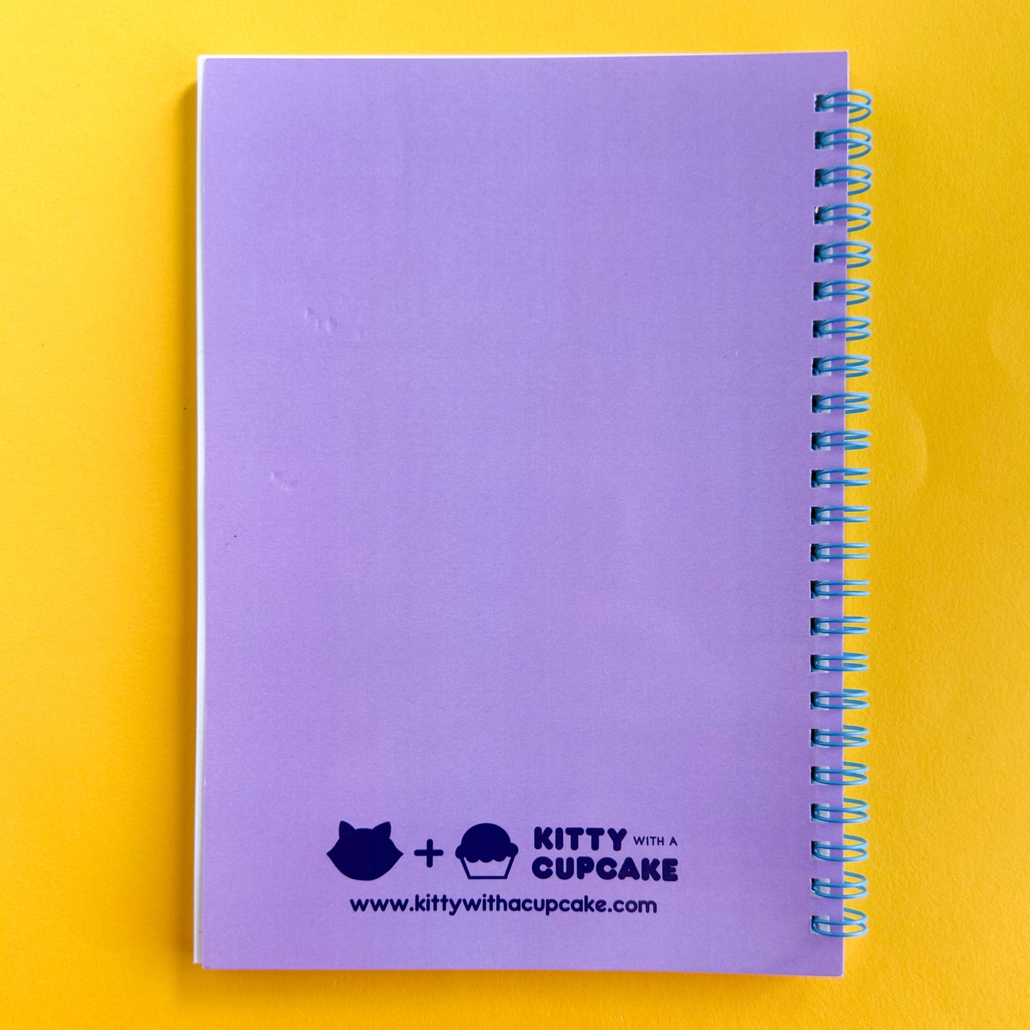 A spiral notebook with a lavender back cover with a Kitty With A Cupcake Logo and www.kittywithacupcake.com printed at the bottom center in dark purple bubble font. The notebook is sitting on a yellow paper background. 