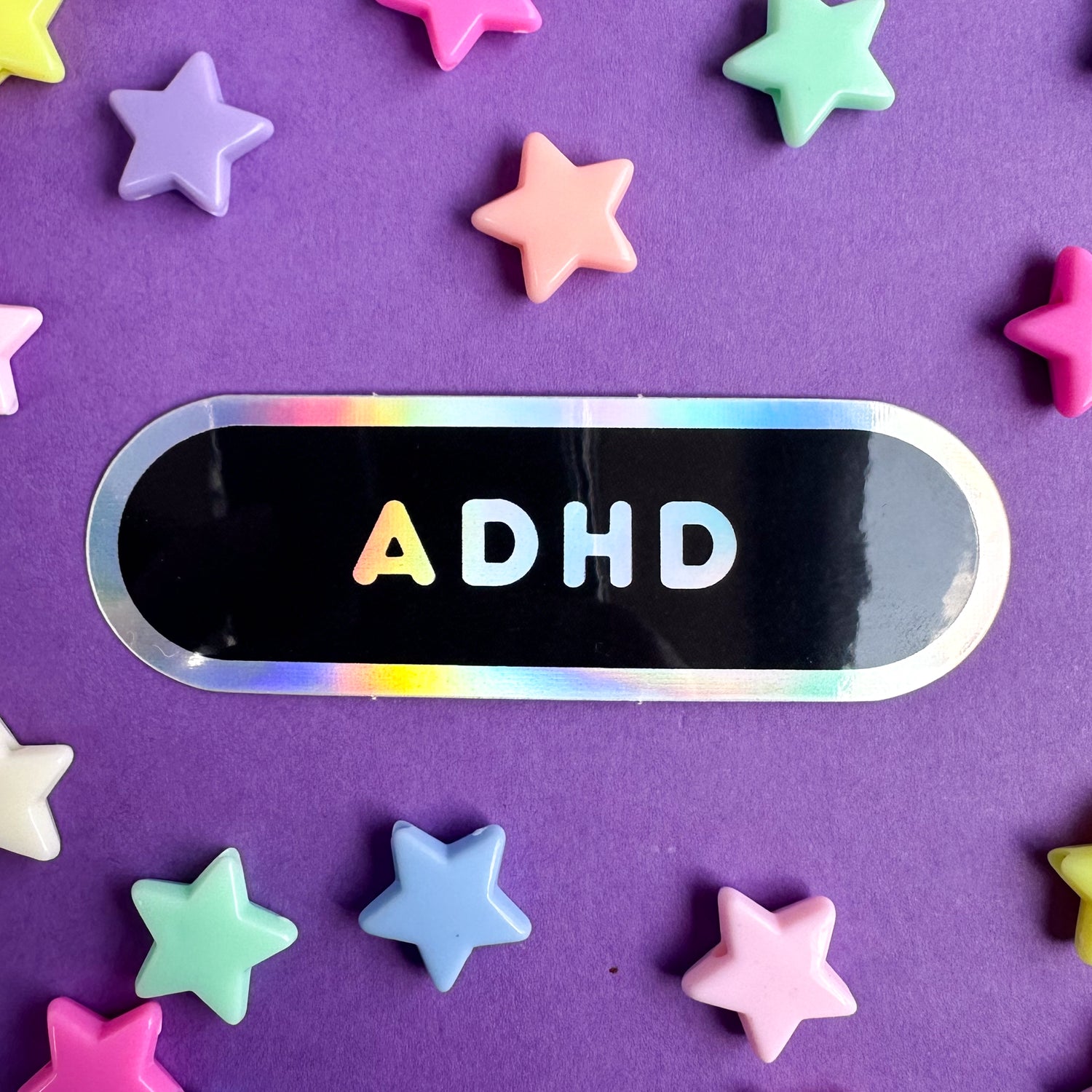 A black bandaid shaped sticker with holographic border and holographic bubble letters reading ADHD on it. 