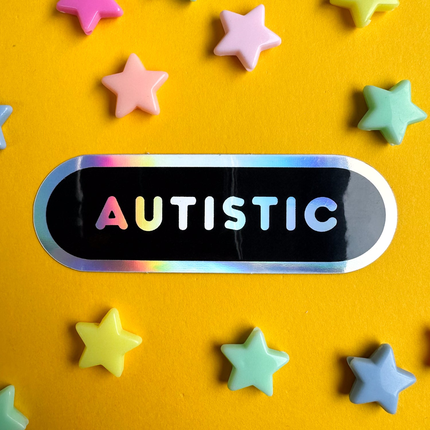 A vinyl sticker in the shape of a black oval with holographic border and holographic bubble letters that spell "Autistic". The sticker is on a yellow piece of paper with pastel star beads around it. 