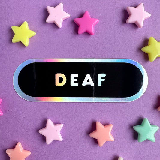 An oval shaped sticker that is mostly black with holographic border and holographic bubble letters that read "Deaf". The sticker is on a purple paper background with pastel plastic star beads scattered around it. 