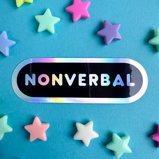 A black capsule shaped sticker with a holographic border and holographic bubble letters. The sticker is on a blue background with pastel plastic star beads around it. 