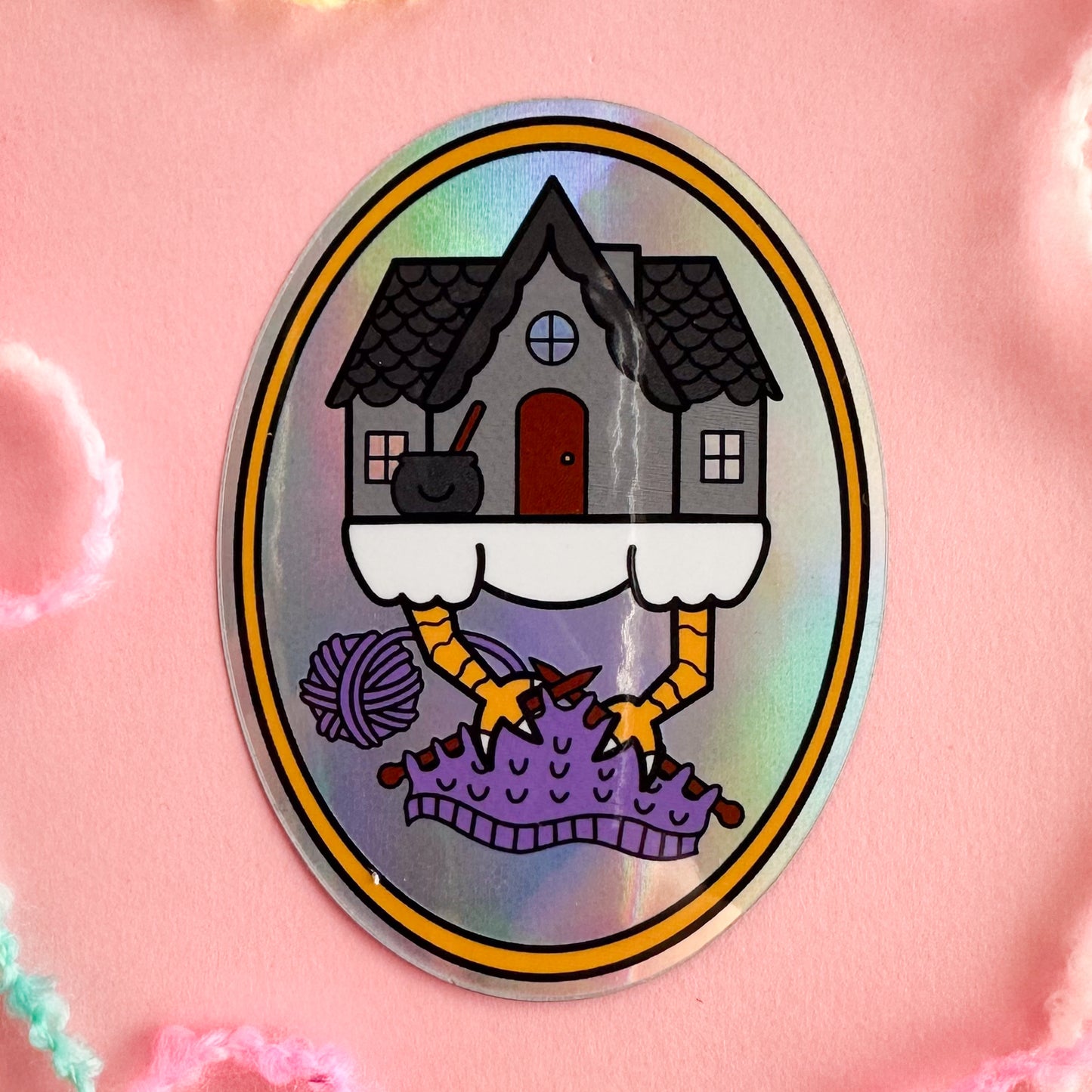 A holographic sticker in the shape of an oval with an illustration of a house with chicken feet knitting on a purple knitting project with it's chicken feet.