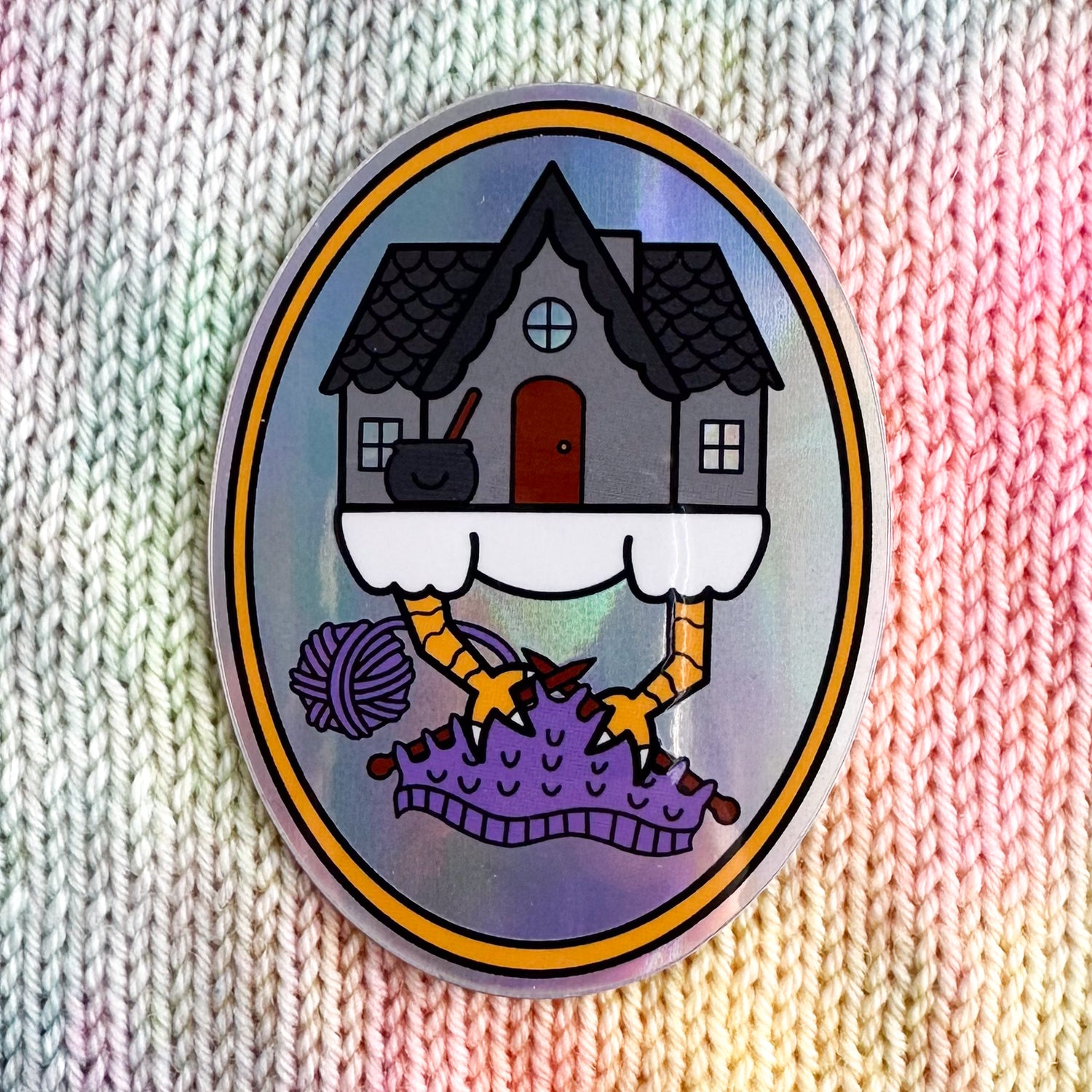 An oval shaped sticker with an illustration of Baba Yaga's house knitting with its chicken feet. The sticker has a holographic background and is sitting on a backdrop of stockinette stitch in pastel colored yarn. 