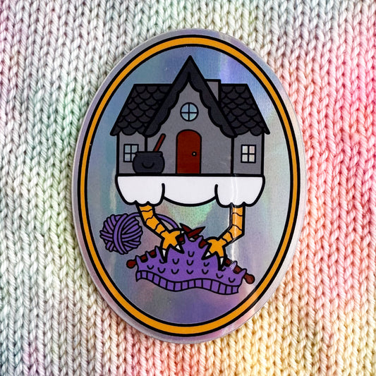 An oval shaped sticker with an illustration of Baba Yaga's house knitting with its chicken feet. The sticker has a holographic background and is sitting on a backdrop of stockinette stitch in pastel colored yarn.