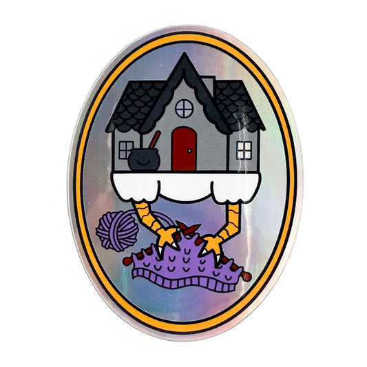 An oval shaped sticker with a cute illustration of Baba Yaga's house knitting on a purple project. The sticker has a holographic effect in the background. 