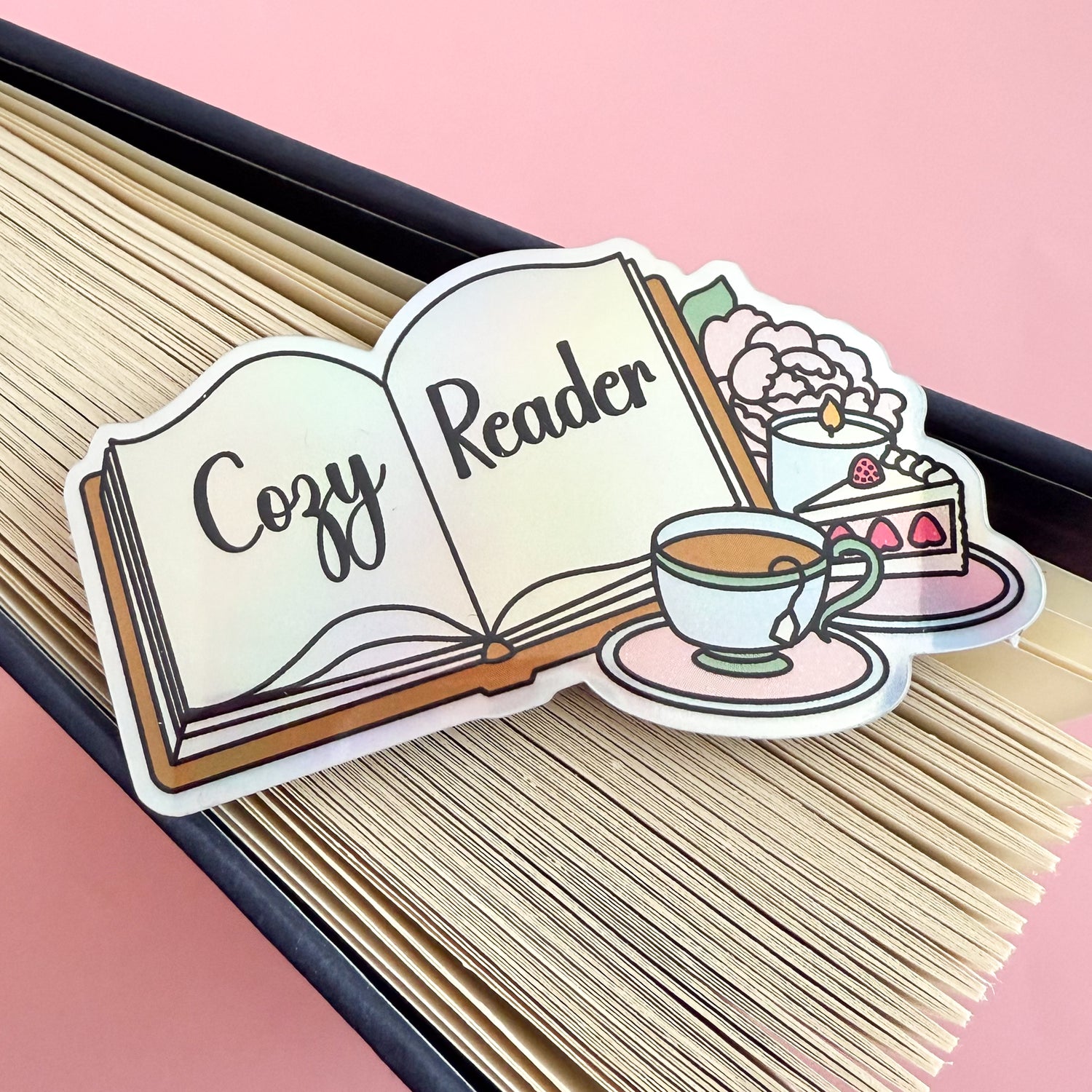 A holographic sticker depicting an open book with “Cozy Reader” written across the pages. Beside the book are a full tea cup on a pink saucer, a slice of strawberry cake, a lit candle, and a baby pink peony with a green leaf. The sticker is displayed and the splayed edge of a book.