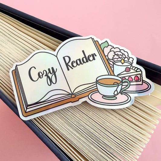 A holographic sticker depicting an open book with “Cozy Reader” written across the pages. Beside the book are a full tea cup on a pink saucer, a slice of strawberry cake, a lit candle, and a baby pink peony with a green leaf. The sticker is displayed and the splayed edge of a book.