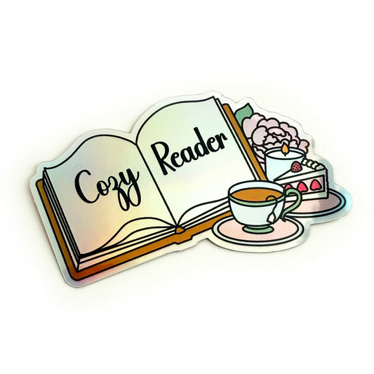 A holographic sticker depicting an open book with “Cozy Reader” written across the pages. Beside the book are a full tea cup on a pink saucer, a slice of strawberry cake, a lit candle, and a baby pink peony with a green leaf.