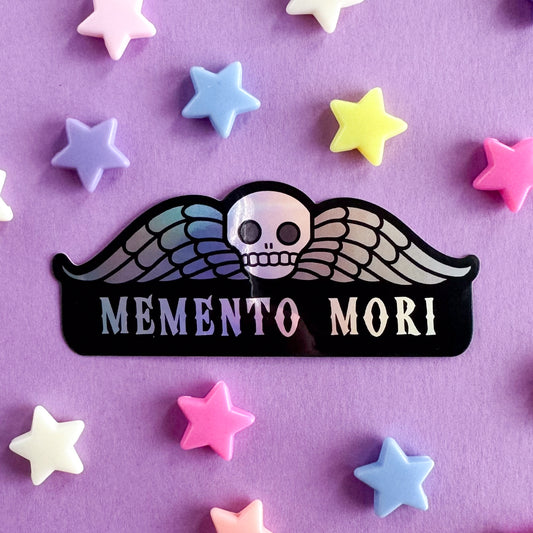 A sticker with an illustration of a deaths head gravestone (a skull with wings) with the words Memento Mori underneath it. The sticker is on a purple background with star shaped beads around it.