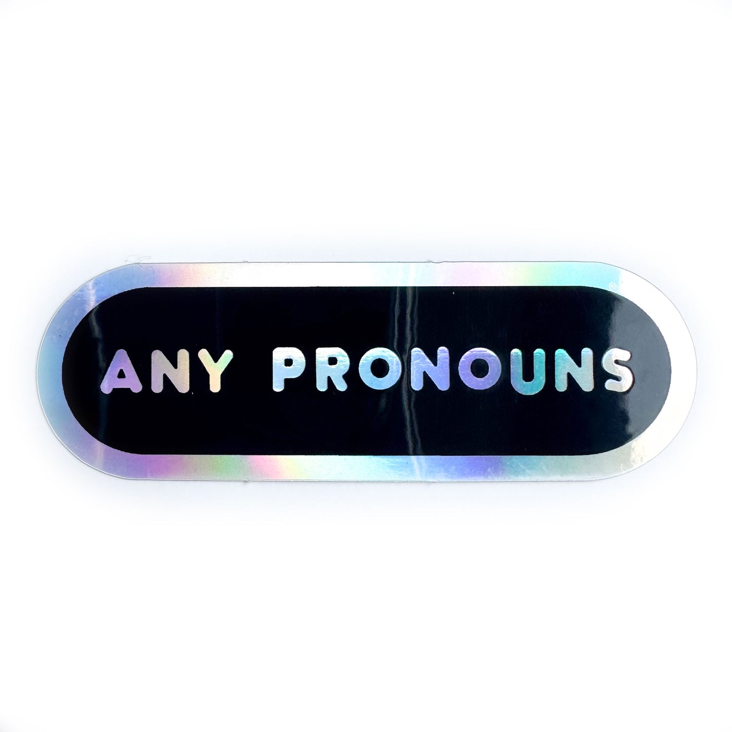 A holographic capsule sticker with a black background with holographic words reading "Any Pronouns"
