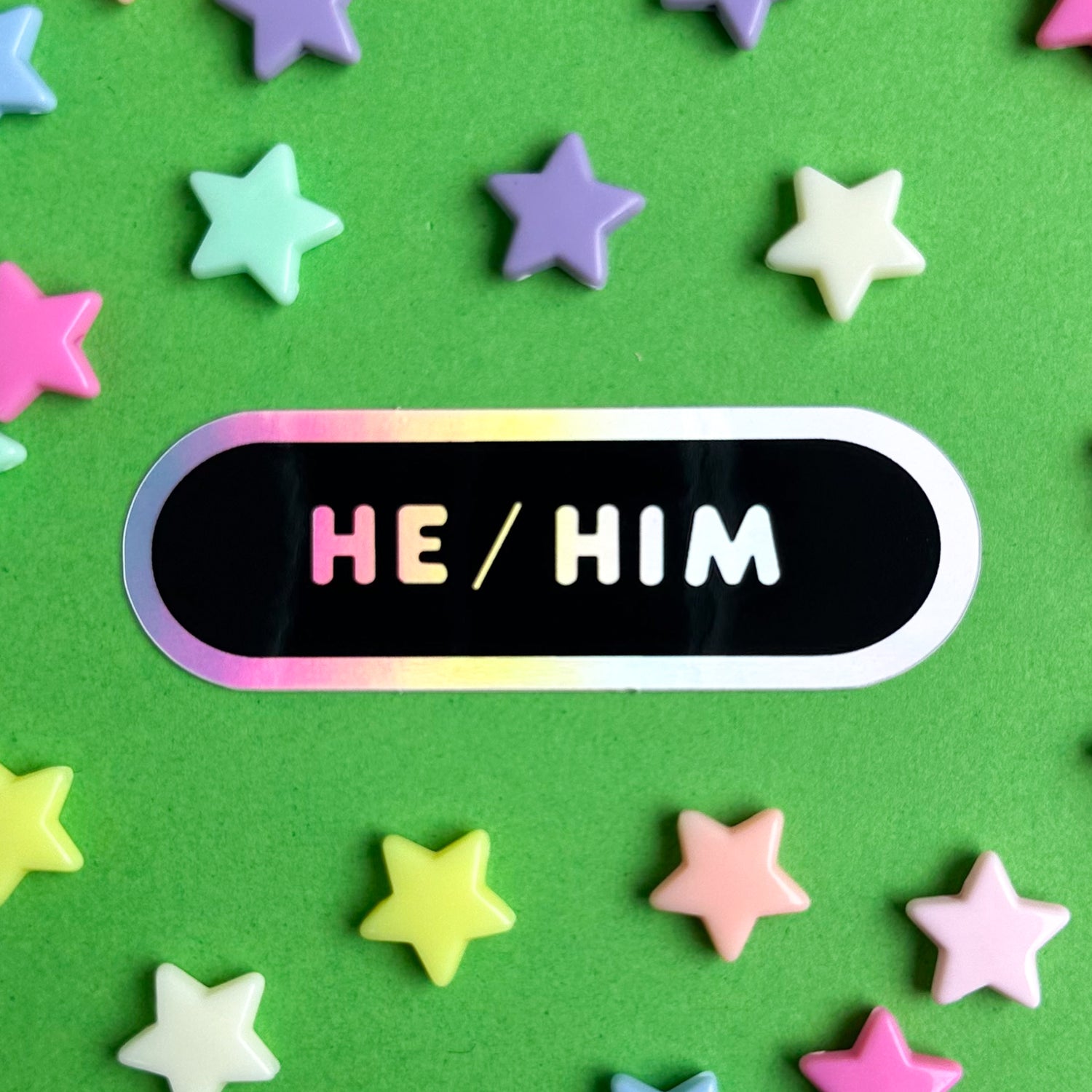 A black oval sticker with the words "He/Him" in holographic bubble letters. The sticker is on a green background with pastel stars around it. 