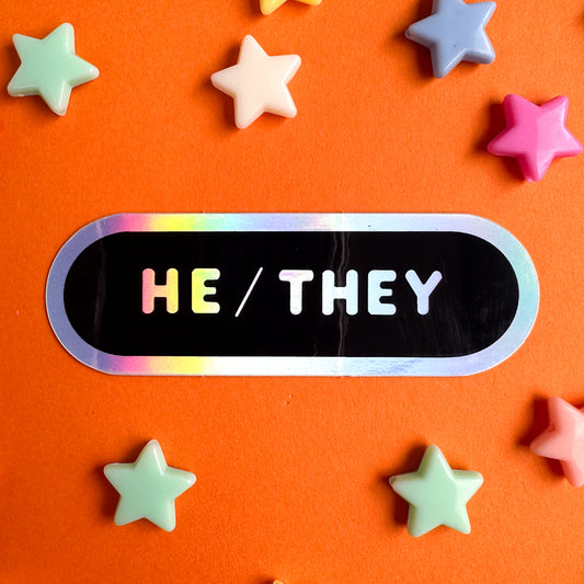 A capsule shaped sticker with holographic text that reads "He/They" on a black background. The sticker is on top of orange paper with star beads around it.