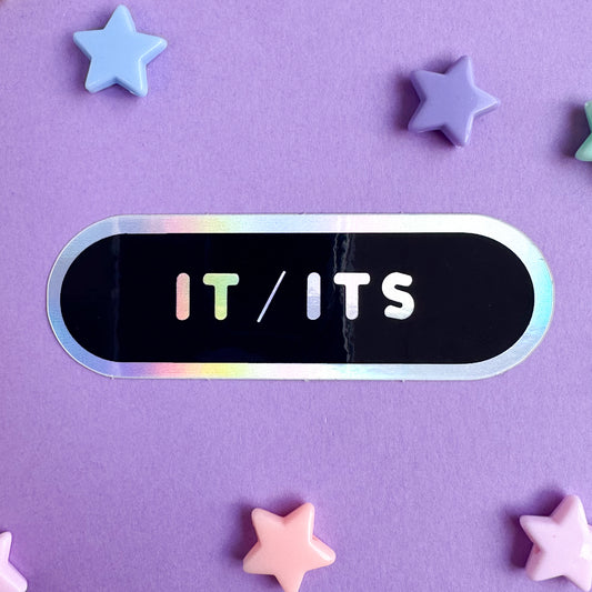 An oval sticker with a black background, holographic border, and holographic words "Its/Its". The sticker is sitting on a purple background with star beads around it.