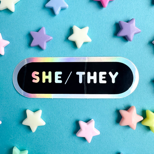 An oval shaped sticker with the words "She/They" in holographic letters on a blackground. The sticker is on a blue piece of paper with star beads around it.