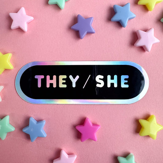 A black oval shaped sticker with a holographic border and "They/She" in holographic bubble letters. The sticker is on a pink background with pastel star beads around it. 