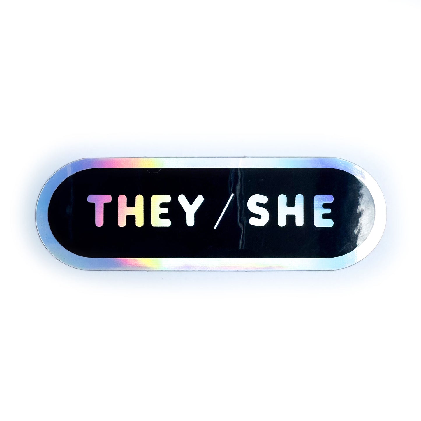 A black capsule shaped sticker with a holographic border and holographic bubble letters that spell the pronouns "They/She".