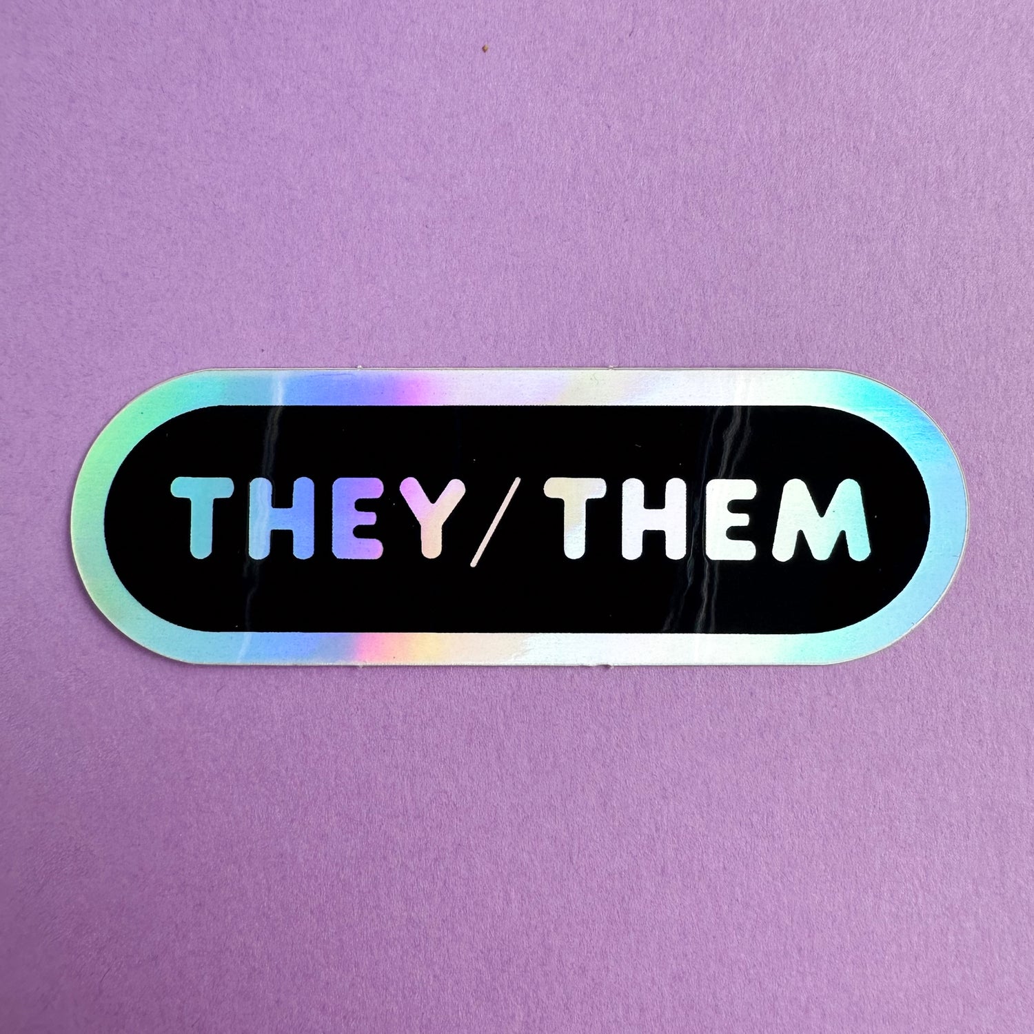An oval shaped sticker with the words "They/Them" in holographic bubble letters on a black background. The sticker is on a purple piece of paper. 