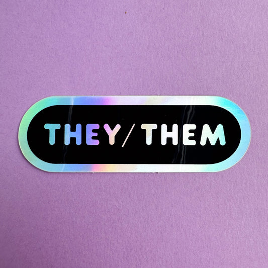 An oval shaped sticker with the words "They/Them" in holographic bubble letters on a black background. The sticker is on a purple piece of paper.