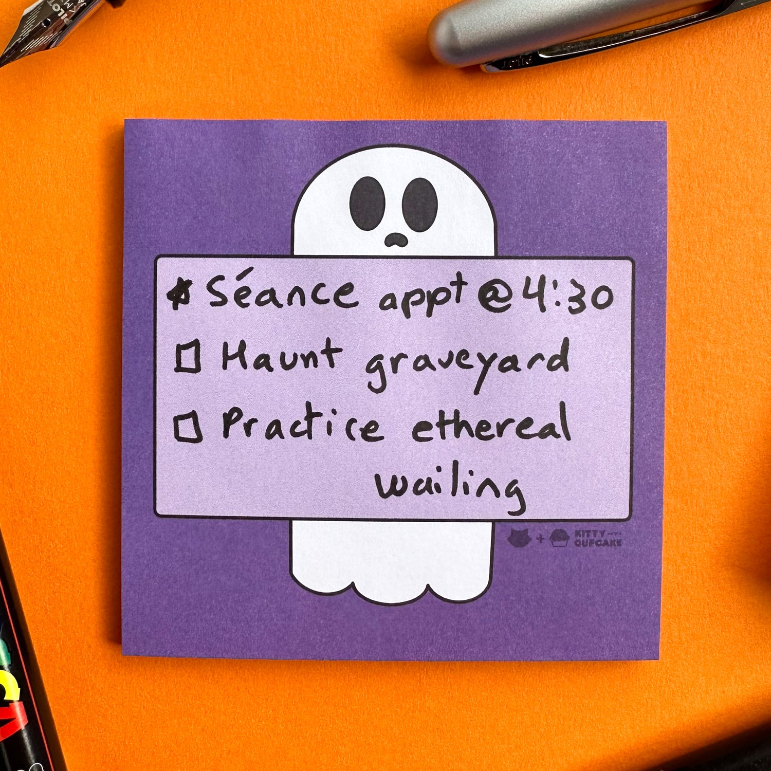 A purple square sticky notepad featuring an illustration of a ghost holding a box with a to-do list written on it. The to-do list reads "Seance appt @ 4:30, Haunt Graveyard,  Practice ethereal wailing". The notepad is on an orange background surrounded by pens.