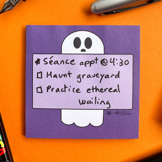 A purple square sticky notepad featuring an illustration of a ghost holding a box with a to-do list written on it. The to-do list reads "Seance appt @ 4:30, Haunt Graveyard, Practice ethereal wailing". The notepad is on an orange background surrounded by pens.