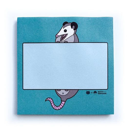 A blue square sticky note pad featuring an illustration of an opossum with its mouth open holding a light blue rectangle to write in. 