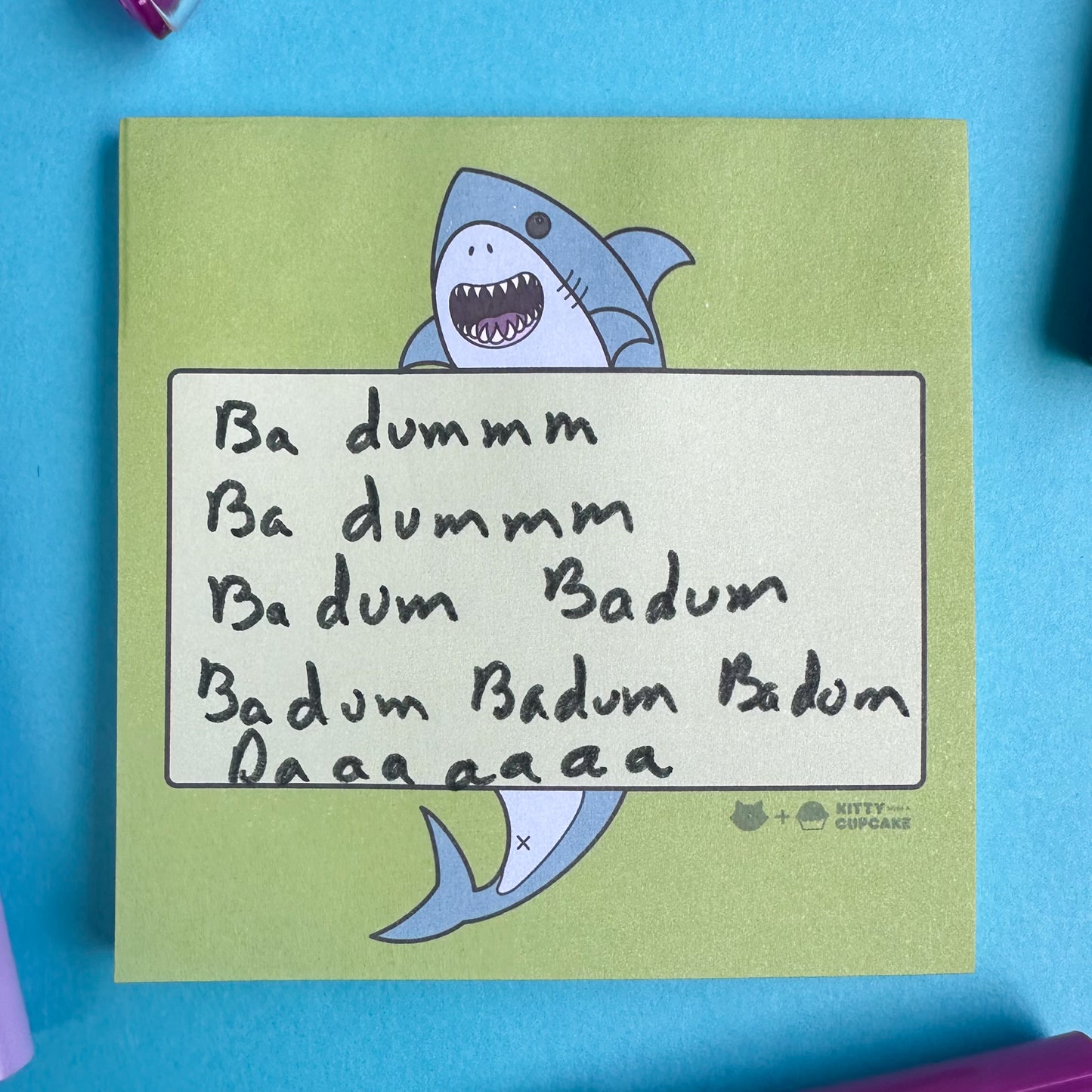 A lime green pad of sticky notes with a cute shark printed on them holding a lighter green box. There is a handwritten note that reads "Da dummm ba dummm bad dum ba dum badum badum badum Daaaaaaaaa" 